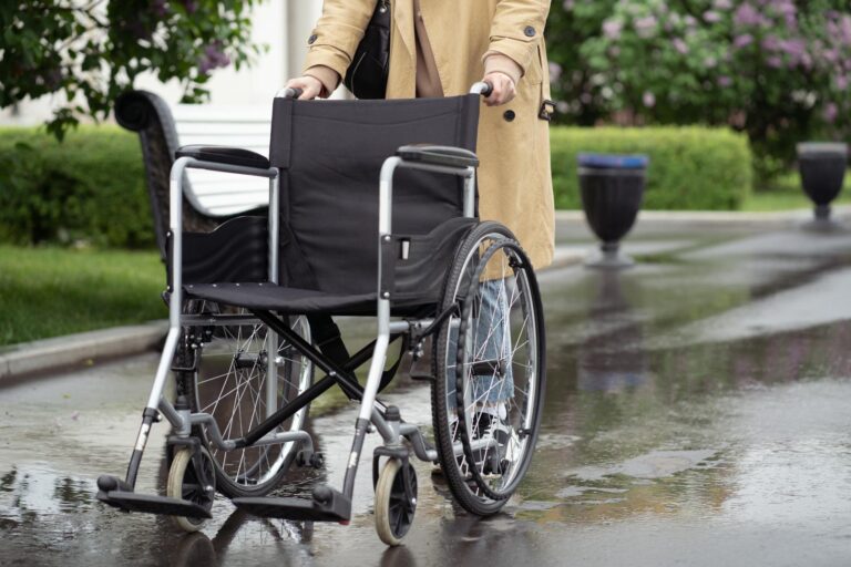 A person in a trench coat pushing an empty wheelchair along a wet outdoor path.