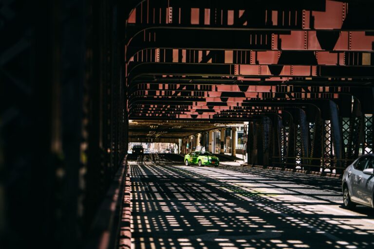 Cars driving under a steel bridge, creating dramatic shadow patterns on the road.